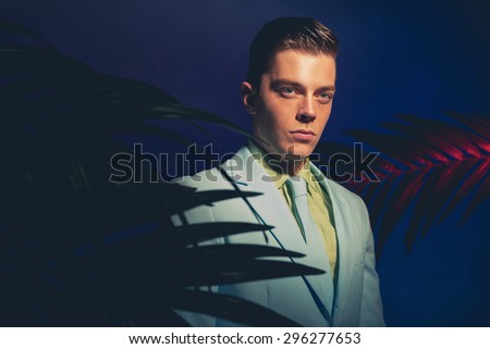 Handsome Man in Formal Wear Standing Between Palm Leaves Against Dark Blue Violet Background.