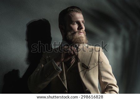 Stern thoughtful bearded man in stylish clothes standing leaning back against a wall looking off to the right of the frame with a serious expression, upper body with shadows