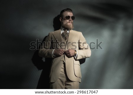 Stylish bearded man wearing trendy sunglasses standing leaning against a wall buttoning his jacket and looking towards a beam of light on the right of the frame