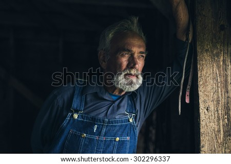 Elderly man in denim dungarees standing staring through a window in an old rustic wooden house or barn with a thoughtful expression