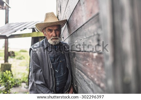 Half Body Shot of a Middle Aged Bearded Male Farmer Wearing Straw Hat and Jacket, Leaning Against Wall of Farmhouse and Looking Straight at the Camera.
