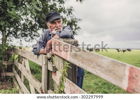Elderly farm worker tending his cattle in the pasture standing leaning on a wooden fence with a serious contemplative expression