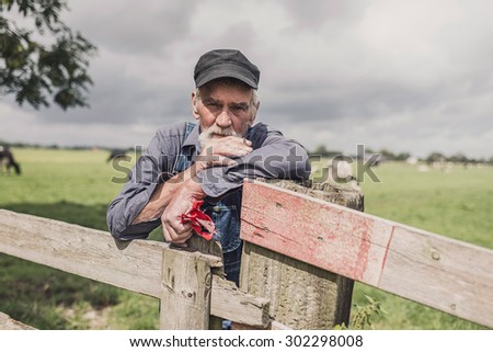 Senior farmer wearing a cloth cap standing smoking in a pasture with a herd of cows leaning on the fence looking at the camera