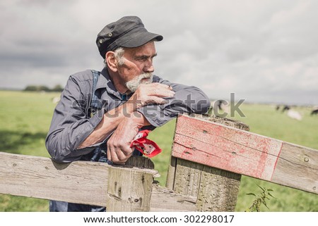 Senior farmer wearing a cloth cap standing smoking in a pasture with a herd of cows leaning on the fence looking at the camera