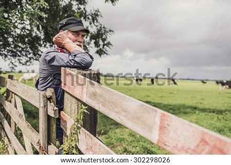 Elderly farm worker tending his cattle in the pasture standing leaning on a wooden fence with a serious contemplative expression