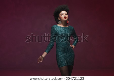 Three-quarter shot of an elegant young woman, in a glittery green dress, looking at the camera. Isolated on maroon background.
