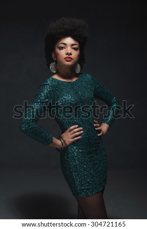 Three-Quarter Shot of an Elegant Young Woman Posing in Sparkling Green Dress with Hands on Waist and Looking at the Camera Against Black Wall Background Inside the Studio.