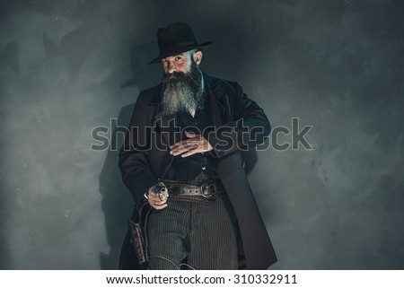Gun shooting vintage crook with long beard in 1900 style clothing against grey wall.