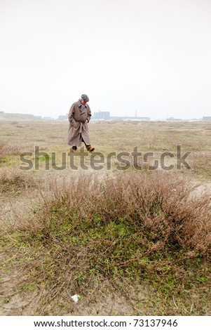 Senior man with raincoat and hat walking on sand with grass. Open space. Lost.