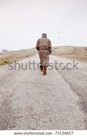 Lonely senior man with raincoat and hat walking on road. Lost.