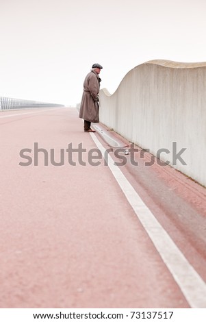 Lost senior man with raincoat and hat walking on empty road on cloudy day.