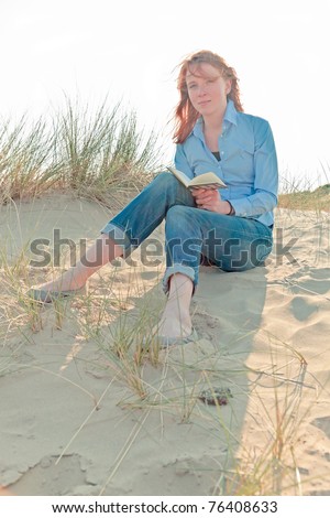 Young woman with red hair sitting on sand dune enjoying nature and reading book.