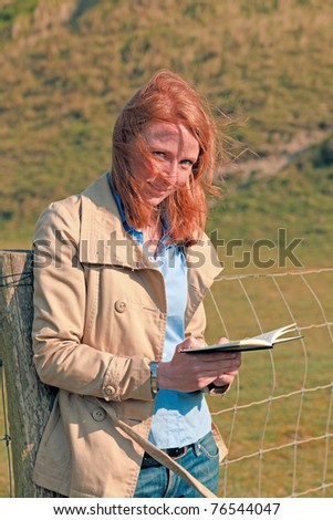 Young woman red hair standing in nature reading book.