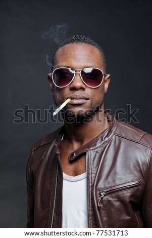 Confident casual young black man wearing brown leather jacket and sunglasses smoking cigarette. Studio portrait against dark background.