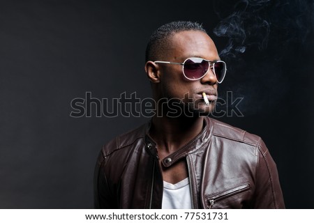 Confident casual young black man wearing brown leather jacket and sunglasses smoking cigarette. Studio portrait against dark background.