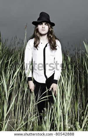 Young man with long brown hair wearing black hat standing in field with long grass. Stormy cloudy sky.