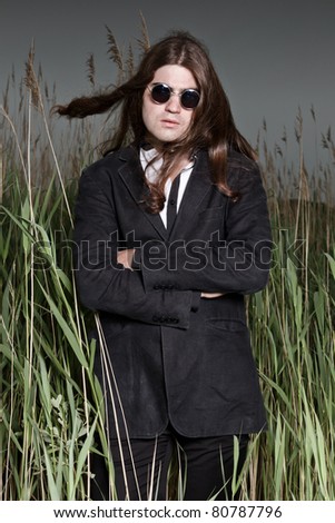 Young man with long brown hair wearing sunglasses and black suit standing in field with long grass. Stormy cloudy sky.