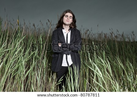 Young man with long brown hair wearing black suit standing in field with long grass. Stormy cloudy sky.