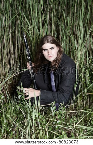 Young man with long brown hair wearing black suit holding clarinet in field of long grass. Stormy cloudy sky.
