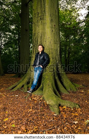 Single casual young man in forest leaning against tree. Short hair wearing jeans and black leather jacket.