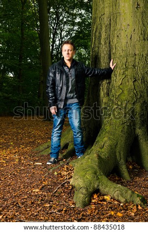 Single casual young man in forest standing near tree. Short hair wearing jeans and black leather jacket.