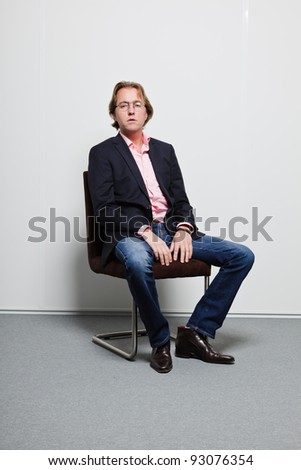 Young business man with blond hair in blue suit and pink shirt wearing glasses sitting on chair in office