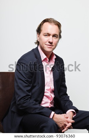 Smiling young business man with blond hair in blue suit and pink shirt sitting on chair isolated on white background