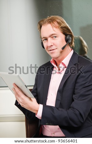 Young business man blond hair with headset and tablet wearing blue suit and pink shirt in front of window.