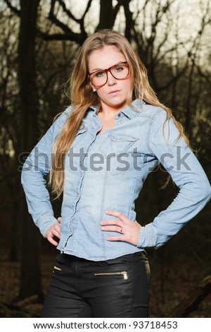 Pretty young woman with long blond hair wearing glasses in winter forest. Wearing blue jeans shirt and black pants.