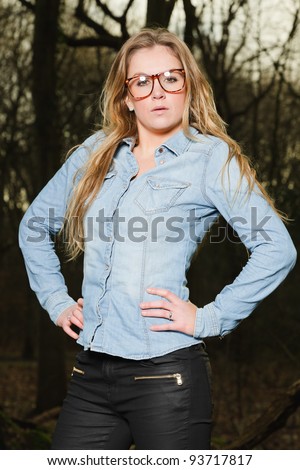Pretty young woman long blond hair wearing glasses in winter forest. Wearing blue jeans shirt and black pants.
