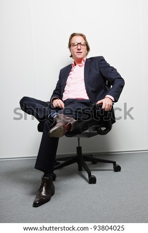 Young business man with blond hair in blue suit and pink shirt sitting on chair in office. Wearing glasses.