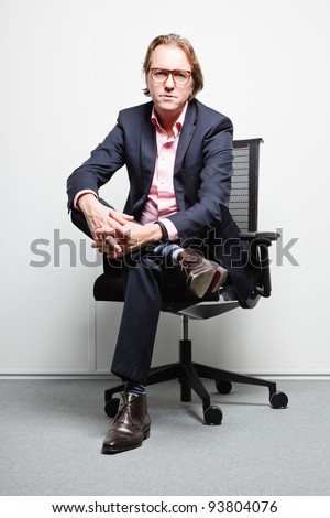 Young business man with blond hair in blue suit and pink shirt sitting on chair in office. Wearing glasses.