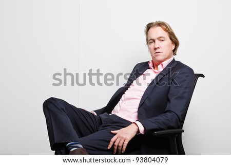 Young business man with blond hair in blue suit and pink shirt sitting on chair in office