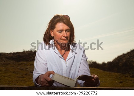 Pretty woman middle aged reading a book and enjoying outdoors. Clear sunny spring day with blue sky.