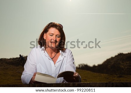 Pretty woman middle aged reading a book and enjoying outdoors. Clear sunny spring day with blue sky.