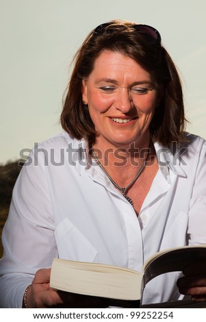 Attractive woman middle aged reading a book and enjoying outdoors. Clear sunny spring day with blue sky.