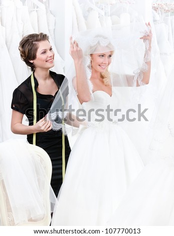 Shop assistant helps to the bride to put the wedding gown on. Bride raises the veil