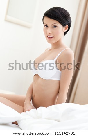 Halfnaked woman in bra sits on the wide bed with white bed linen