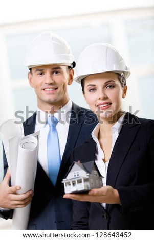 Two engineers in white hard hats keeping blueprints and model house
