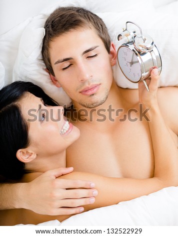 Close up of couple lying in bed. Woman holds alarm clock near the ear of man, top view
