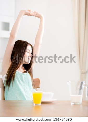 Woman stretches herself sitting at the kitchen table with breakfast and glass of orange juice