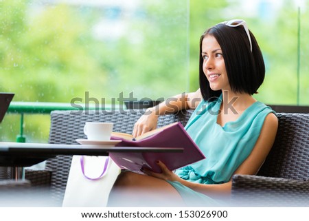 Girl wearing blue dress and sunglasses sits at the table of the bar and reads book with cup of tea