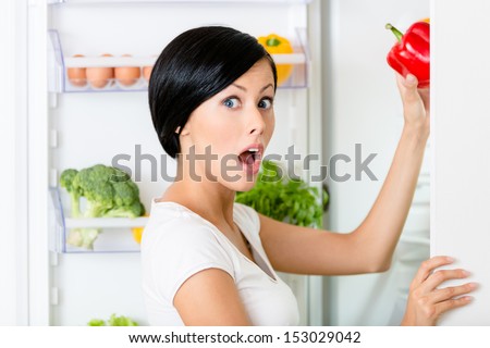 Young woman takes red pepper from the opened fridge full of vegetables and fruit. Concept of healthy and dieting food