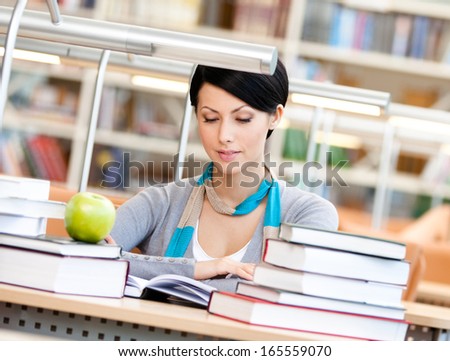 Woman with green apple surrounded with piles of books reads sitting at the table at the library. Information overload