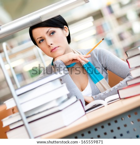 Pretty female student studies with piles of books sitting at the desk at the reading hall