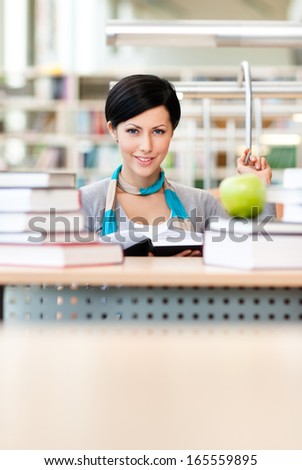 Female student with green apple studies sitting at the desk at the reading hall of the library. Information concept