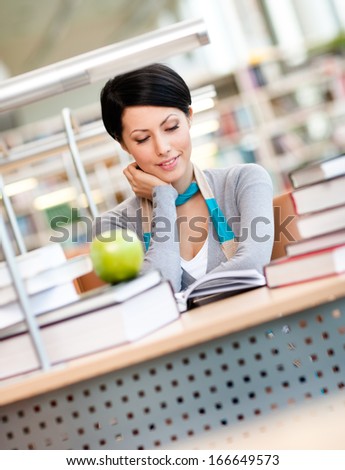 Female student with green apple studies sitting at the desk at the reading hall of the library. Academic achievement
