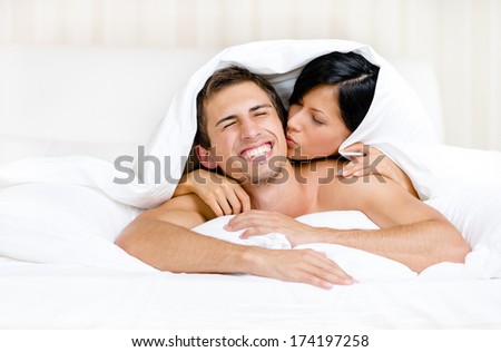 Close up of couple playing in bed. Covered with blanket woman lying on the back of the man kisses him