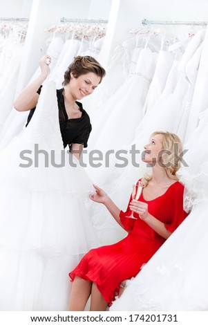 Shop assistant suggests another dress to the bride while she is drinking champagne