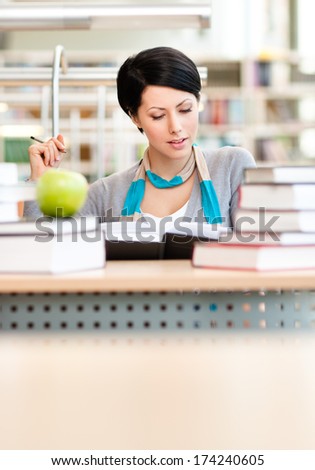 Interested female student with green apple studies sitting at the desk at the reading hall of the library. Knowledge concept